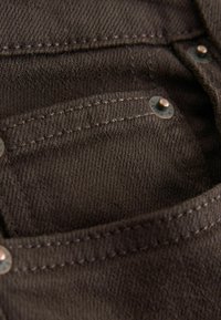 Primo piano di un tessuto in denim marrone che presenta una piccola tasca con rivetti in metallo e cuciture visibili, evidenziando texture e elementi di design durevoli.