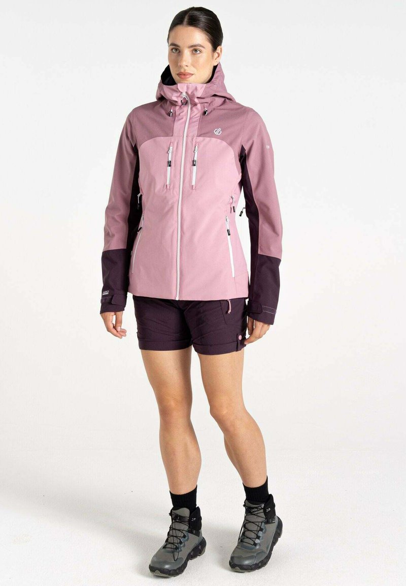 Rosa und lila wasserdichte Jacke mit Kapuze, ausgestattet mit zwei Reißverschluss-Brusttaschen und elastischen Bündchen, getragen mit dunklen Shorts und Wanderstiefeln.