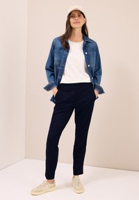 Donna con un cappello bianco, giacca di jeans, t-shirt bianca, pantaloni blu navy e sneakers beige, in piedi con le mani in tasca davanti a un muro beige.