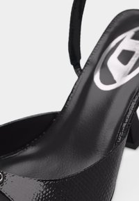 Μαύρο παπούτσι slingback από δέρμα με λουστρίνι, τακούνι block, υφή στην επιφάνεια και ορατό λογότυπο της μάρκας στο εσωτερικό της σόλας.