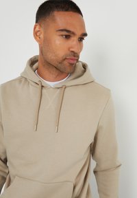 Sudadera beige con bolsillo frontal, capucha con cordón y pequeño logo. Textura suave y lisa con un diseño minimalista y color neutro.