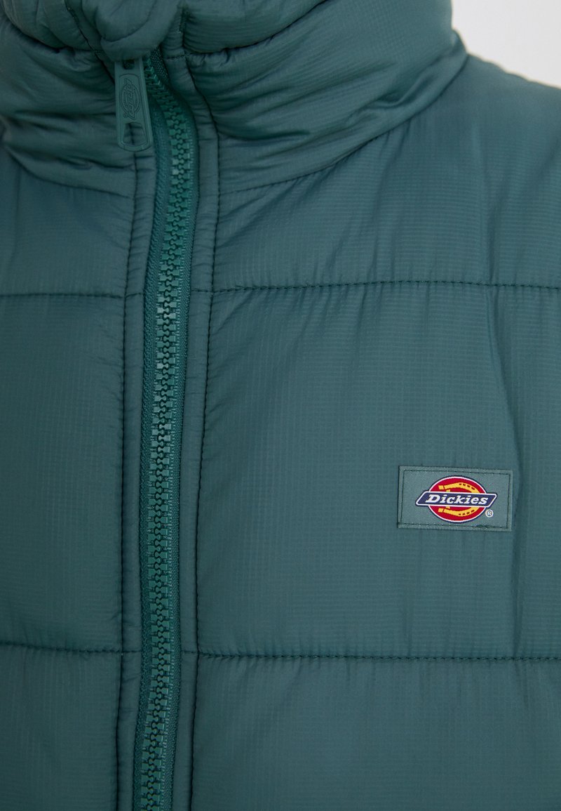 Grüne Pufferjacke mit hohem Kragen, einem zentralen Reißverschluss und einem Logo-Patch auf der linken Seite. Das Material hat eine glatte Textur mit genähten Paneelen.