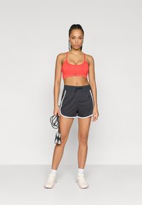 Κόκκινο αθλητικό σουτιέν με λεπτές τιράντες, συνδυασμένο με μαύρα σορτς με λευκές πλευρικές ρίγες, κρατώντας ένα σχοινάκι, με φόντο άσπρο.