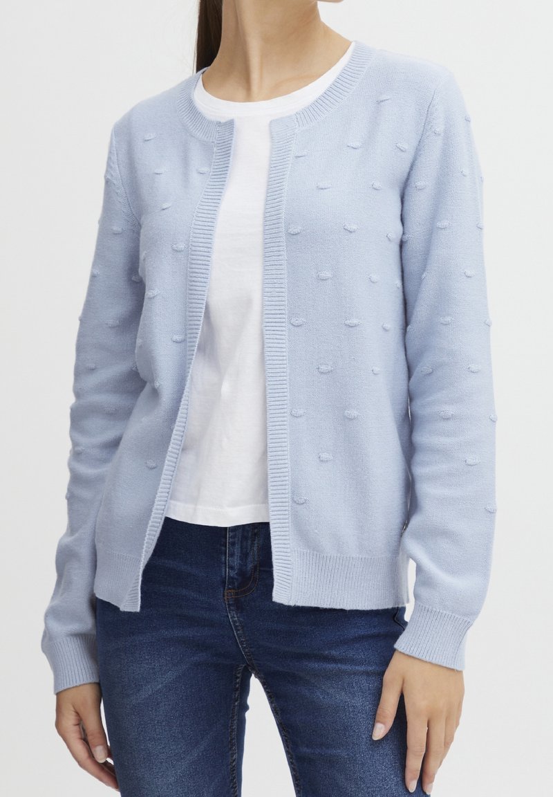 Femme portant un cardigan en maille bleu clair avec des points texturés sur un t-shirt blanc et un jean bleu foncé, debout devant un fond uni.