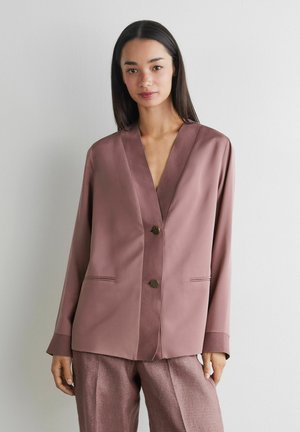 Kobieta o długich ciemnych włosach, ubrana w mauve satynową marynarkę i pasujące spodnie, stoi na tle jednolitej jasnej ściany.