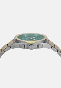 Reloj de hombre con esfera verde, caja hexagonal de acero inoxidable con acentos dorados, pulsera de eslabones metálicos y dos botones en el lateral.