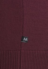 Pull en tricot marron avec une texture côtelée, une couture texturée et une petite étiquette noire avec un 'M' blanc à l'ourlet.