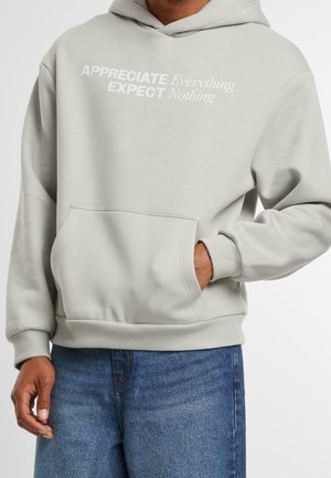 Mężczyzna w jasnoszarej bluzie z kapturem z przednią kieszenią i napisem "Doceniaj wszystko, nie oczekuj niczego" w połączeniu z niebieskimi dżinsami.