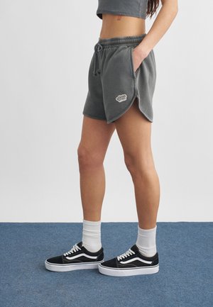 Person iført mørkegrå shorts med snøre og sidespalte, hvide sokker og sorte sneakers med lavt skaft, stående på blåt gulv.
