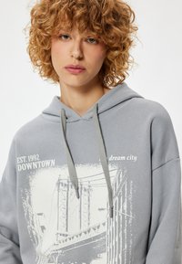 Grijze hoodie met een grafische print van een brug en stadslandschap. Voorzien van een voorzak, verstelbare koorden en zachte textuur.