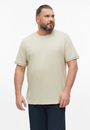 Behaarde man met naar achter getrokken haar draagt een lichtbeige T-shirt met korte mouwen en een donkere broek, staand tegen een effen witte achtergrond.