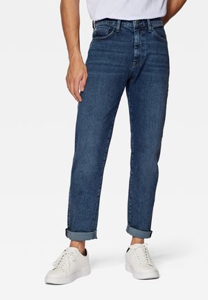 Mann, der dunkelblaue Jeans mit Umschlag und weiße Sneaker trägt, steht mit einer Hand entspannt an der Seite.