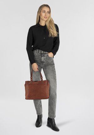 Borsa tote in pelle marrone con hardware della tracolla, tenuta da una persona in una maglietta nera e jeans a vita alta grigi, con stivali neri con tacco.