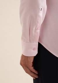 Polsino di camicia rosa chiaro con una texture liscia, caratterizzato da due bottoni rosa e un piccolo design ricamato nero. La camicia è infilata in pantaloni scuri.
