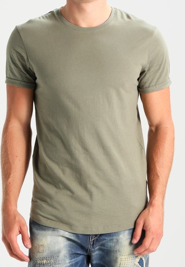 Homme portant un t-shirt uni vert olive à manches courtes et un jean bleu usé, debout devant un fond uni.