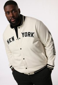 Krämfärgad varsityjacka med "NEW YORK" i svarta bokstäver, randiga manschetter och en slät textur. Matchas med svarta rutiga byxor.