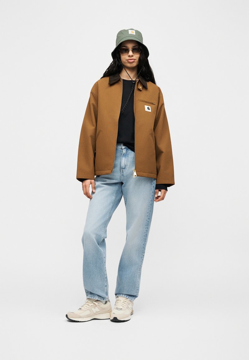 Vrouw met een bruine jas, zwart shirt, lichtblauwe jeans, beige sneakers, groene bucket hat en zonnebril, staand tegen een effen achtergrond.