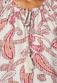 Blusa leve e estampada com decote ajustável, base predominantemente branca com padrões paisley em vermelho, rosa e amarelo, e um tassel branco.
