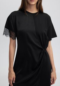 Robe noire à manches courtes. Une manche est ornée de dentelle. Design enveloppant avec un détail de nœud à la taille. Texture de tissu lisse et douce.