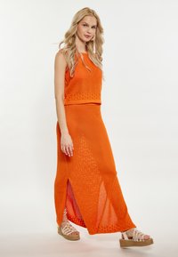 Oranje gebreid twee-delig set bestaande uit een mouwloos topje en een lange rok met zijsplits. De outfit heeft een gestructureerd patroon en is gecombineerd met wiggen sandalen.