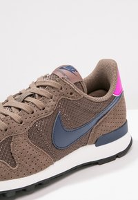 Baskets Nike marron en daim perforé, panneaux en mesh, logo Swoosh bleu foncé, semelle intermédiaire blanche, semelle extérieure noire et languette de talon rose vif.