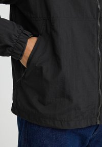 Veste coupe-vent légère noire avec des poignets élastiques, des poches latérales zippées et un tissu lisse et texturé. Portée sur un jean bleu.