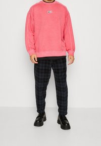 Sudadera rosa con cuello de canalé y parche con logotipo, combinada con pantalones de pijama negros a cuadros y zapatos negros gruesos.