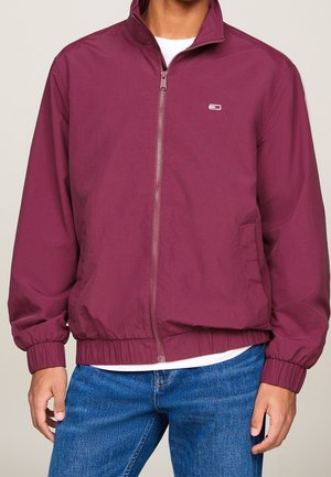 Mand iført en vinrød lynlåsoverdel med elastiske manchetter og kant, lille logo på brystet, kombineret med blå jeans og hvid undertrøje.