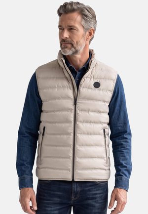 Mand iført let beige quiltet vest over blå denimskjorte og mørke jeans, kigger til siden mod en ensfarvet baggrund.