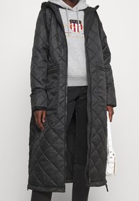 Persona che indossa un lungo cappotto nero imbottito sopra un hoodie grigio con logo, pantaloni neri e tiene una piccola borsa bianca al suo fianco.