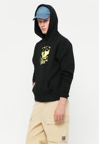 Black hoodie with a front graphic design in yellow, featuring a skull. The model pairs it with beige cargo pants and a denim cap.
