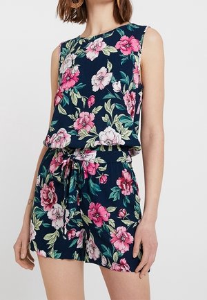 Mouwloze marineblauwe romper met roze en witte bloemenprint, een gestrikte tailleband en een ontspannen pasvorm, gedragen door een staande vrouw.