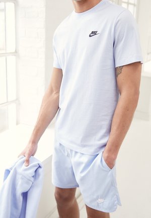 Mężczyzna w jasnoniebieskiej koszulce i spodenkach Nike, trzymający pasującą jasnoniebieską kurtkę, stoi przy oknie w jasnym pokoju.