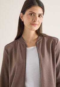 Femme aux longs cheveux bruns et taches de rousseur, portant une chemise blanche et une veste marron zippée, regardant légèrement de côté avec une expression neutre.