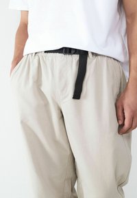 Persoon in losse beige broek met een zwarte riem en een wit shirt, met de handen in de zakken, staande tegen een effen achtergrond.
