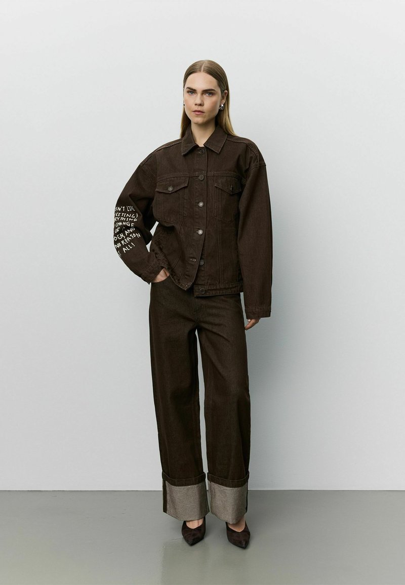 Femme debout portant une veste oversize marron foncé avec un texte blanc sur la manche, un pantalon large retroussé et des chaussures pointues foncées sur un fond uni.