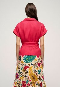 Blusa rossa a maniche corte con vita sottolineata da una cintura e colletto, abbinata a una maxi gonna colorata e decorata con motivi floreali e paisley.