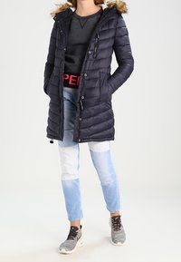 Doudoune marine avec capuche en fourrure, dotée d'une fermeture éclair et de boutons, de poches latérales et d'une texture matelassée. Portée sur un haut gris et un jean.