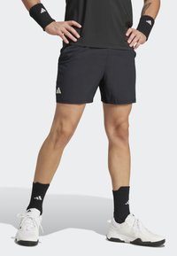 Schwarze Sportshorts mit elastischem Bund, strukturiertem Stoff und einem kleinen weißen Logo auf der linken Seite. Weiße Sneaker und schwarze Knö socken.