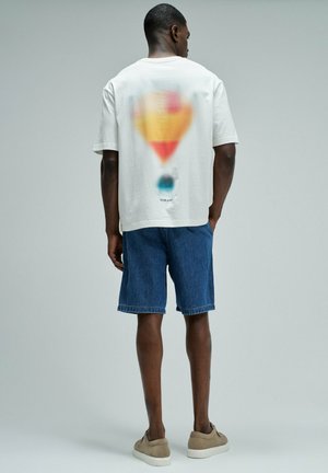 Homme portant des baskets beige, un short en jean bleu et un T-shirt blanc oversize avec un graphisme flou de montgolfière colorée dans le dos.