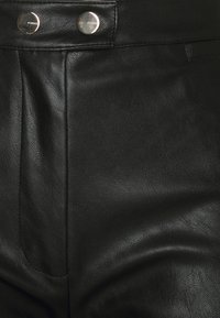 Gros plan sur un pantalon en cuir noir texturé avec deux boutons métalliques gravés « PINKO » sur la ceinture.