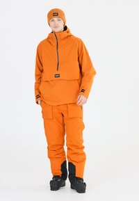 Orange wasserdichte Oberbekleidung mit Kapuze, Reißverschlusstasche und großer Fronttasche. Kombiniert mit passenden Hosen und schwarzen Stiefeln.