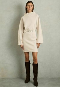 Rochie bej din jerseu, cu guler înalt, mâneci lungi și talie ajustată, asortată cu cizme din suede maro, până la genunchi, și o curea albă.