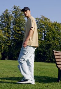 Camiseta oversized beige, pantalones vaqueros holgados azul claro con bolsillos, gorra negra y zapatillas negras, situados en un parque con hierba y árboles.