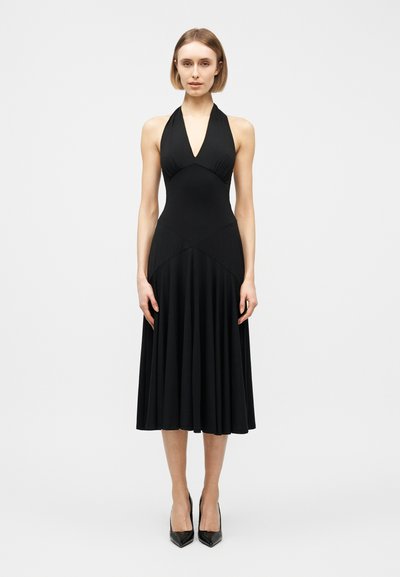 Femme debout portant une robe noire sans manches à col licou avec une jupe midi plissée et des escarpins noirs pointus sur un fond blanc uni.