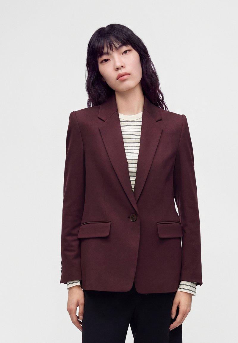 Femme aux longs cheveux noirs porte un blazer bordeaux foncé sur une chemise rayée blanche et noire, sur un fond clair uni.