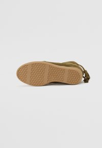 Bottines en suède vert olive avec une semelle en caoutchouc marron texturée présentant un motif géométrique et des franges décoratives à l'arrière.