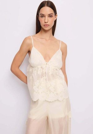 Giovane donna con capelli castani lisci che indossa un top in pizzo bianco con spalline e pantaloni larghi coordinati, posando davanti a uno sfondo chiaro e semplice.