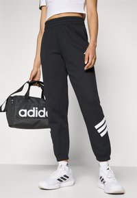 Pantaloni della tuta neri con vita elastica, dettagli a tre strisce bianche lungo le gambe, abbinati a una borsa da palestra nera con un logo bianco.
