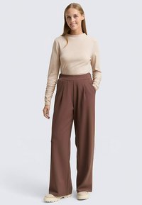 Beige top met lange mouwen en bruine wijde broek met plooien aan de voorkant. Model staat met handen in de zakken, draagt beige chunky schoenen.